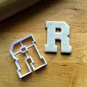 May include: White plastic cookie cutter in the shape of the letter R. The letter is in a bold, blocky font style. A white cookie in the shape of the letter R is next to the cutter.