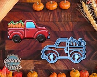May include: A red truck cookie decorated with orange pumpkins in the bed and a light blue window, alongside a light blue plastic truck-shaped cookie cutter. The cookie features a smooth finish, while the cutter shows a detailed outline design.