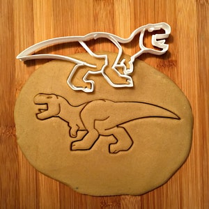 May include: A white plastic Tyrannosaurus Rex cookie cutter sits atop a cut-out cookie of the same shape. The cookie is light brown, and the background is a wooden surface.