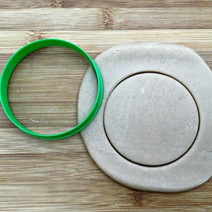 May include: A green plastic cookie cutter is placed on a wooden cutting board next to a circle of beige dough that has been cut out with the cookie cutter.