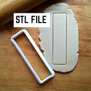 May include: A white plastic cookie cutter in the shape of a rectangle. The cookie cutter is on a wooden surface with a piece of dough that has been cut out with the cookie cutter.