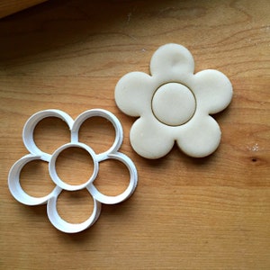 Puede incluir: Un cortador de galletas de pl&aacute;stico blanco con forma de flor de seis p&eacute;talos y un c&iacute;rculo central. Junto a &eacute;l hay una galleta que se ha cortado con el cortador de galletas.