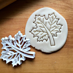 May include: White plastic cookie cutter in the shape of a detailed maple leaf. The cutter is shown on a wooden surface next to a piece of dough with the leaf design pressed into it.