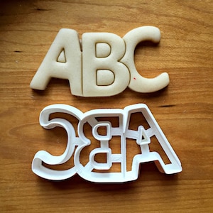 May include: A cookie cutter and a baked cookie, both in the shape of the letters "ABC". The cookie is light brown, and the cutter is white. The cookie is on a wooden surface.