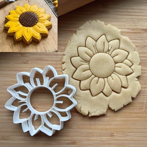 May include: A white plastic sunflower cookie cutter next to a piece of rolled dough with a sunflower impression. An inset shows a finished cookie decorated with yellow icing petals and a brown center on a wooden surface.