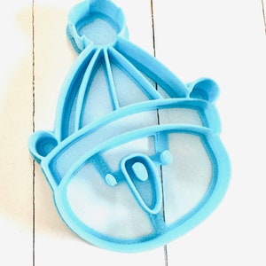 May include: A light blue plastic cookie cutter shaped like a snowman wearing a hat. The snowman has a round head with two eyes and a mouth.