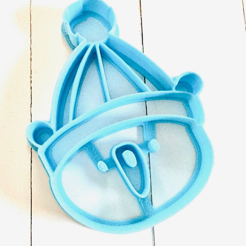 May include: A light blue plastic cookie cutter shaped like a snowman wearing a hat. The snowman has a round head with two eyes and a mouth.