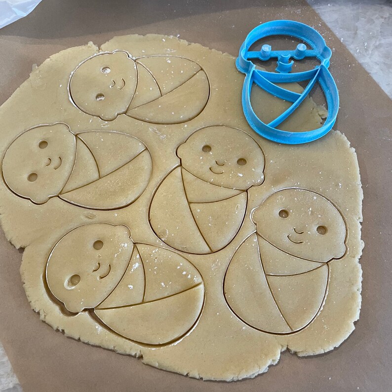 May include: A close-up of a sheet of cookie dough with five baby-shaped cookie cutters pressed into it. The cookie cutters are blue plastic and have a simple design.