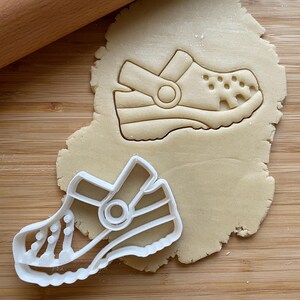 May include: White plastic cookie cutter shaped like a sandal with a strap and a round hole. The cutter is on top of a piece of rolled out cookie dough.