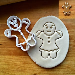 K&ouml;nnte beinhalten: Ein wei&szlig;er Ausstechform aus Kunststoff in Form eines Lebkuchenmanns neben einem St&uuml;ck ausgerolltem Teig mit demselben eingedr&uuml;ckten Motiv. Ein kleiner fertiger Lebkuchen mit wei&szlig;er Glasur ist in der oberen Ecke zu sehen.