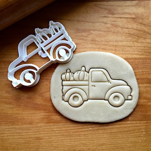 May include: A white plastic cookie cutter in the shape of a pickup truck with a bed full of pumpkins. The cutter is on top of a piece of unbaked cookie dough.