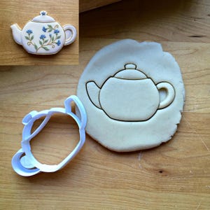 May include: A white plastic teapot-shaped cookie cutter next to a circular piece of dough with a teapot impression. An inset shows a finished cookie decorated with white icing, blue cornflower patterns, and gold trim on the teapot edges.