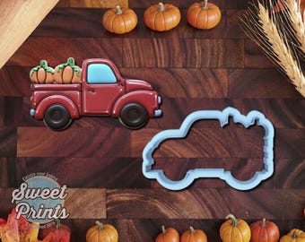 May include: A red truck cookie decorated with two orange pumpkins in the bed, featuring a light blue window and black wheels, alongside a matching light blue plastic truck-shaped cookie cutter on a dark wooden surface with small pumpkins.