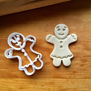 K&ouml;nnte beinhalten: Ein wei&szlig;er Ausstechform aus Kunststoff in Form eines Lebkuchenm&auml;nnchens neben einem passenden hellbeigen Keks. Die Form zeigt den Umriss einer Figur mit Kleid, gewellten &Auml;rmeln und Saum, w&auml;hrend der Keks gepr&auml;gte Gesichtsz&uuml;ge aufweist.