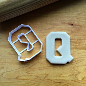 May include: Two white plastic cookie cutters shaped like the letters Q and R. The cutters are on a wooden surface.