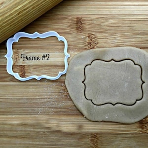 May include: A white plastic cookie cutter in the shape of a frame with the text "Frame #2" inside. The cookie cutter is on a wooden surface next to a piece of dough that has been cut out with the cookie cutter.