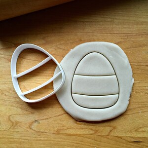 May include: A white plastic Easter egg cookie cutter beside a piece of dough with an egg shape cut into it. The egg shape has two horizontal lines. The background is a wooden surface.
