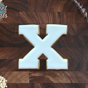 May include: A letter X shaped cookie with smooth white icing on top. The cookie has a golden brown edge and is positioned centrally. The logo for Sweet Prints Established 2015 is visible in the top left corner of the image.