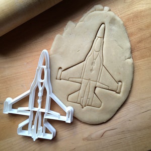 May include: A white plastic cookie cutter shaped like a fighter jet, with a brown cookie dough cutout of the same shape.