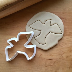 Peut inclure: Un emporte-pièce en plastique blanc en forme de colombe sur une surface en bois à côté d'une pâte découpée en forme de colombe.