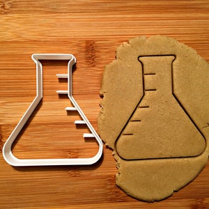 May include: A white plastic cookie cutter shaped like a beaker with a graduated scale on the side. The cutter is next to a round piece of brown cookie dough with the same shape cut out.