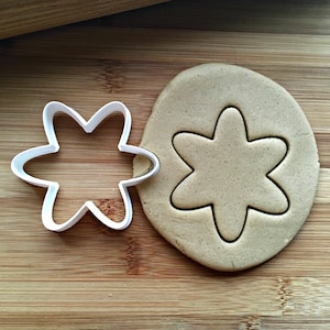 May include: A white plastic flower-shaped cookie cutter next to a piece of cut dough. The dough is light brown and the cutter is white. The background is a wooden cutting board.