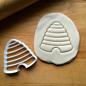 May include: A white plastic beehive cookie cutter with a white dough imprint of the beehive. The cookie cutter is on a wooden surface.
