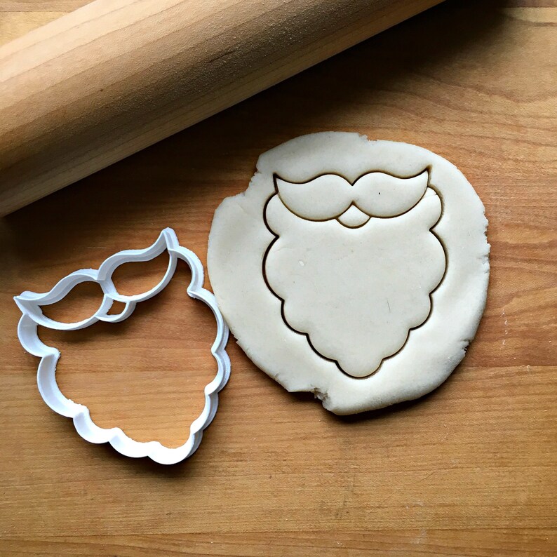 May include: A white plastic cookie cutter in the shape of Santa Claus's beard and mustache sits next to a piece of dough with the same shape cut out. A wooden rolling pin is in the background. The items are on a wooden surface.