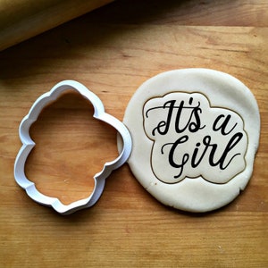May include: A white plastic cookie cutter in a cloud shape next to a round piece of dough with the words "It's a Girl" in black script. The dough is imprinted with the cutter's shape. The background is a wooden surface.