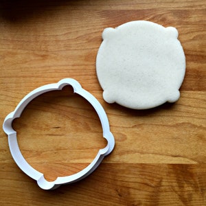 May include: A white plastic cookie cutter in the shape of a rounded square with four rounded corners, next to a cookie cut in the same shape. The cookie and cutter are on a wooden surface.