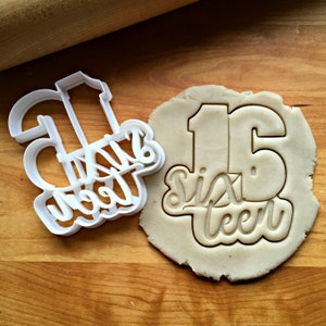May include: A white plastic cookie cutter in the shape of the number "16" with the words "Sixteen" in cursive. Next to it is a piece of dough with the "16" and "Sixteen" imprint. The items are on a wooden surface.
