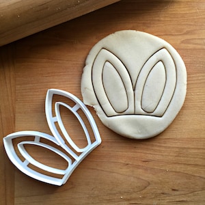 May include: A white plastic bunny ear-shaped cookie cutter next to a round piece of dough with the same bunny ear shape pressed into it. A wooden rolling pin is in the background. The items are on a wooden surface.