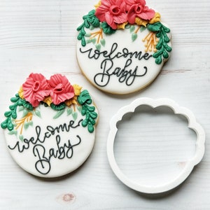 May include: Two round sugar cookies decorated with green leaves, pink flowers, and black icing that says "Welcome Baby". A white plastic cookie cutter is also shown.