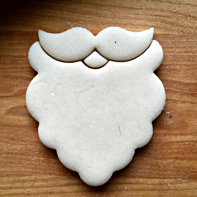 May include: A light-colored, Santa Claus beard-shaped cookie cutter. The cutter features a mustache and beard design, perfect for holiday baking. The cutter is made of a light-colored material and sits on a wooden surface.