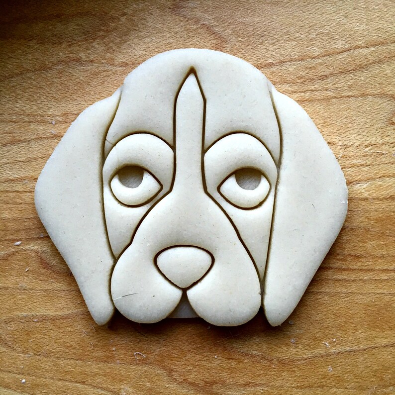 May include: A light-colored dog-shaped cookie cutter. The cutter features a detailed outline of a dog's face, including eyes, nose, and ears. The cutter is made of a light-colored material and is set against a wooden surface.