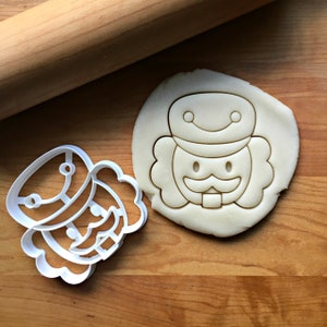May include: White plastic cookie cutter in the shape of a nutcracker. The cutter is on a wooden surface next to a piece of cookie dough.