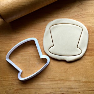 May include: A white plastic top hat cookie cutter next to a piece of dough with a top hat shape cut out. A wooden rolling pin is in the background. The items are on a light brown wooden surface.
