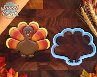 May include: A turkey-shaped cookie decorated with brown, orange, and yellow icing, featuring white eyes and a red wattle, positioned next to a light blue plastic turkey-shaped cookie cutter. The text Sweet Prints is visible in the corner.