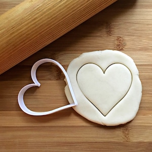 May include: A white heart-shaped cookie cutter on a wooden surface with a rolling pin and a piece of dough with a heart cut out.