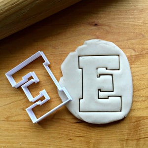 May include: A white plastic cookie cutter in the shape of the letter E. The cutter is on a wooden surface next to a piece of dough with the letter E cut out.