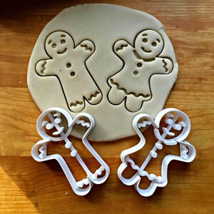 May include: Two white plastic gingerbread cookie cutters shaped like a person and a person wearing a dress. The cutters are positioned below a circular piece of rolled-out beige dough that features two embossed gingerbread figure impressions.