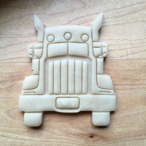 May include: A white cookie cutter shaped like a semi-truck. The cookie cutter has a detailed design with a grill, headlights, and a bumper.