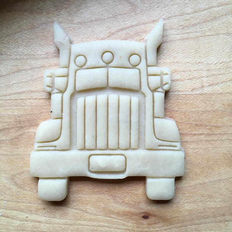 May include: A white cookie cutter shaped like a semi-truck. The cookie cutter has a detailed design with a grill, headlights, and a bumper.