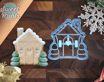 May include: A light blue plastic house-shaped cookie cutter alongside a decorated gingerbread house cookie featuring a white roof, a central heart-shaped door, and two green pine trees on either side, with the Sweet Prints logo visible.