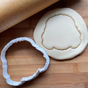 May include: A white plastic cookie cutter in the shape of a cloud, with a piece of cookie dough on a wooden cutting board, and a wooden rolling pin in the background.