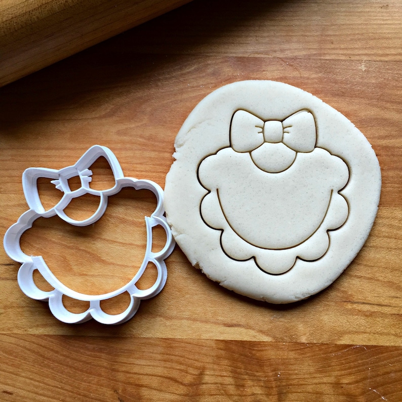 May include: A white plastic cookie cutter in the shape of a bib with a bow on top. The cutter is shown on a wooden surface next to a piece of dough that has been cut out with the cutter.