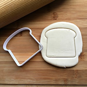 May include: A white plastic cookie cutter shaped like a slice of bread on a wooden surface. A piece of white dough is being cut out with the cookie cutter.