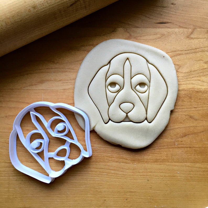 May include: A white plastic dog-shaped cookie cutter next to a cookie with the same dog face design pressed into it. The cookie is on a wooden surface, with a rolling pin in the background.