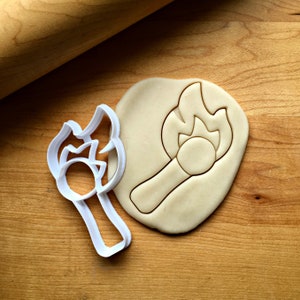 May include: A white plastic cookie cutter in the shape of a lit match, next to a piece of dough with the match imprint. The dough is light brown, and the background is a wooden surface.