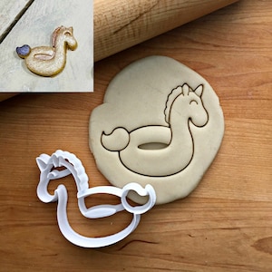 Peut inclure: Un emporte-pièce en plastique blanc en forme de cheval à bascule, à côté d'un biscuit découpé et d'un biscuit fini. Le biscuit fini est doré avec une queue violette. L'ensemble est sur une surface en bois.
